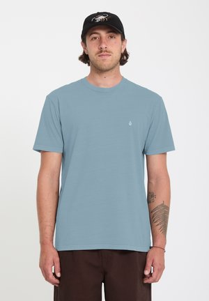 Homme portant un t-shirt à manches courtes bleu clair, une casquette noire avec un logo blanc, et un pantalon marron foncé, debout devant un fond blanc uni.