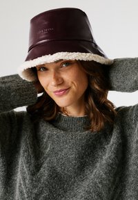 Young woman with freckles wearing a maroon bucket hat with white fleece lining and a gray fuzzy sweater, smiling with hands behind head.