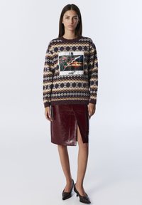 Woman wearing patterned sweater with a floral and fire print, burgundy leather skirt with front slit, and black heels standing against white background.