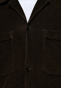 Blazer en velours côtelé marron foncé avec deux poches avant à rabat et des boutons noirs sur une chemise blanche.