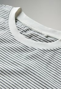 Medvilniniai marškinėliai su apvalia apykakle, plonais juodais dryžiais ant baltos fono. Minkšta tekstūra, matomas siuvimas palei apykaklę.