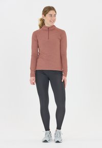 Ung kvinde står og smiler, iført en langærmet støvet rosa zip-up trøje, sorte leggings og lysegrå sneakers, på hvid baggrund.