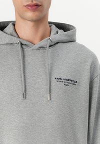 Felpa grigia in cotone con cappuccio e cordino, con logo e testo ricamati in blu navy. Vestibilità standard con una finitura morbida e texturizzata.