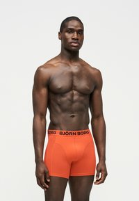 Μυώδης άντρας στέκεται μπροστά σε απλό φόντο, φορώντας πορτοκαλί μπόξερ Björn Borg με μαύρη ζώνη μέσης και κείμενο.