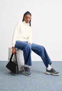 Hvit langermet fleecejakke med glidelås, blå vide jeans, grå og grønne sneakers, holder en svart håndveske. Sitter på et sølvfarget metallobjekt.