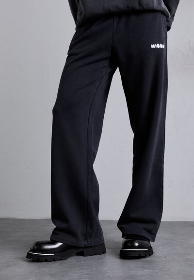 Sorte brede sweatpants i blødt materiale med en hvid logo-detalje på hoften, parret med kraftige sorte sko.