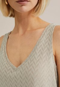 Valkoinen harmaa hihaton toppi, jossa on V-pääntie ja tekstuurinen herringbone-kuosi, pehmeä neulosmateriaali ja näkyvät ompelu yksityiskohdat.