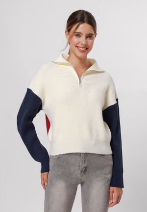 Pull en tricot avec demi-fermeture éclair, présentant un corps crème, des manches bleu marine et rouge, une texture côtelée et un ourlet ajusté.