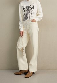 Vit sweatshirt med svart grafisk design, krämfärgade vida byxor och bruna mönstrade stövlar. Håller en liten beiget handväska.