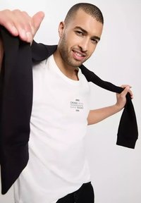 Biała koszulka z czarnym logo i napisem; model trzyma czarną bluzę na ramieniu; gładki materiał, luźny krój.