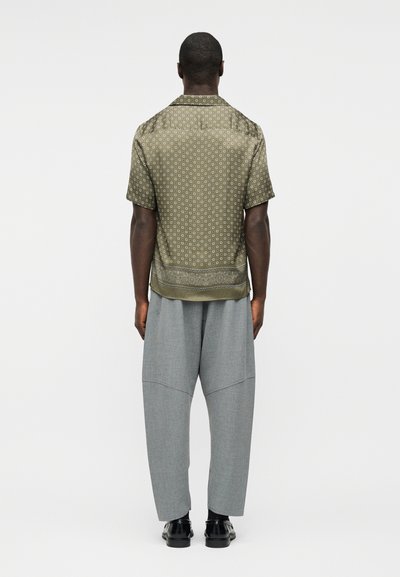 Άνδρας που φαίνεται από πίσω, φοράει πουκάμισο με κοντά μανίκια με ελιά πράσινο μοτίβο, φαρδιά γκρι παντελόνια, μαύρες κάλτσες και μαύρα παπούτσια.