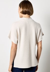 Helles beige gestricktes Oberteil mit einem hohen Kragen, kurzen Ärmeln und einem strukturierten Muster. Der Saum ist hinten leicht verlängert und wird mit dunkelblauen Jeans kombiniert.