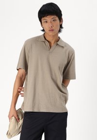 Beige kortermet polo med teksturert mønster, åpen krage og avslappet passform, kombinert med mørke bukser og en lys farget veske.