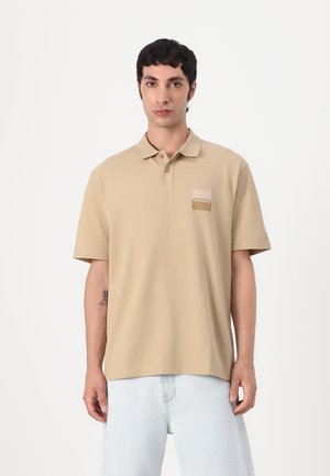 Beige polo-shirt med blød tekstur, med et lille mærke på venstre bryst og korte ærmer. Bæres med lyseblå jeans.