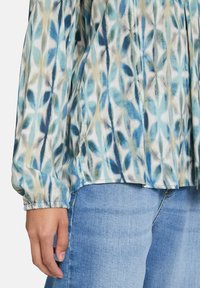Personne portant un blouse à manches longues à motif feuillu bleu avec des poignets élastiques et un jean en denim bleu clair, main détendue sur le côté.