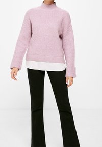 Frau in einem helllila gerippten Pullover mit umgeschlagenen Ärmeln über einem weißen Hemd, kombiniert mit schwarzen Schlaghosen, steht vor einem weißen Hintergrund.