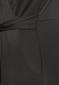 Čierny overal s prekrývajúcim dizajnom, so zaväzovaním v páse, hladkou textúrou a jemným leskom. Obsahuje detail bočných vreciek.