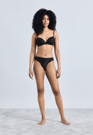 Reggiseno in pizzo nero e perizoma abbinato con bordi smerlati, caratterizzati da spalline sottili e una texture liscia. La modella è in piedi a piedi nudi su uno sfondo chiaro.