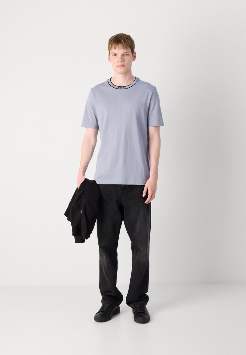 T-shirt grigio chiaro a maniche corte con un collo a coste nero, abbinato a jeans neri e sneakers nere, il tutto completato da una giacca nera.