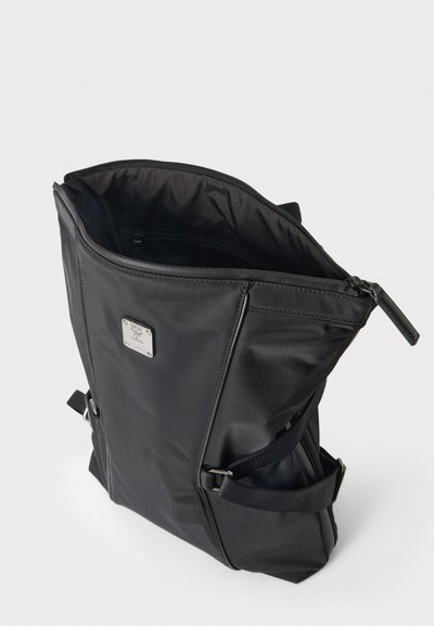 Sac cabas noir avec fermeture éclair ouverte, plaque logo en métal à l'avant, et double poignées, présenté debout sur une surface blanche.