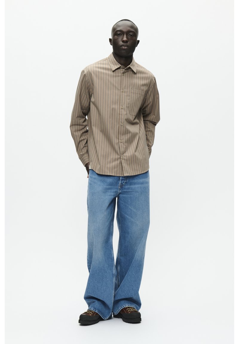 Maglietta a maniche lunghe a strisce marrone chiaro con colletto abbottonato, abbinata a jeans in denim blu larghi. L'outfit presenta un design e una texture minimalista.