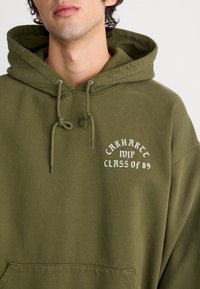 Oliwkowa bluza z kapturem, z sznurkami, z przednią kieszenią kangurką oraz białym napisem "CARHARTT CLASS OF 89" na klatce piersiowej. Miękka struktura materiału.