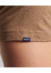 Närbild av brun tygkant med en liten marinblå etikett som läser "SUPERDRY," bredvid bar hud med synliga ljusa hår.