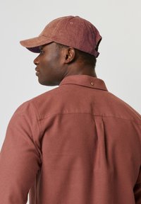 Chemise en velours côtelé bordeaux avec un col à boutons, associée à une casquette texturée mauve avec une visière incurvée et une sangle ajustable à l'arrière.