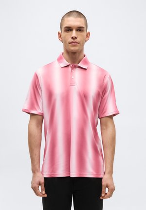 Jeune homme aux cheveux courts portant un polo rayé verticalement rose et blanc et un pantalon noir, debout devant un fond uni.