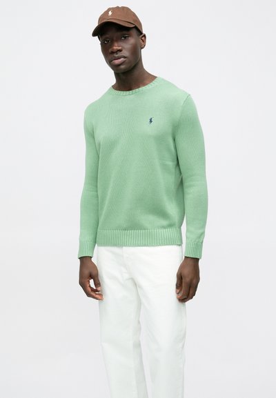 Zeleni pleteni pulover z rebrastimi manšetami in robom, z majhnim mornarsko modrim logotipom. Kombiniran z belimi hlačami, model nosi rjavo kapo.