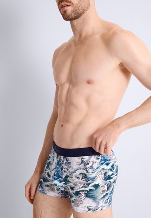Homme torse nu portant un boxer à motifs avec un design océan et figures, posant avec une main ajustant la taille.