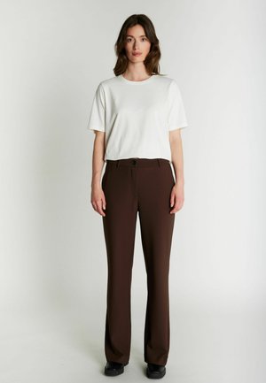 Femme debout face à l’avant, portant un t-shirt blanc uni à manches courtes, un pantalon droit marron foncé et des chaussures noires sur un fond neutre.