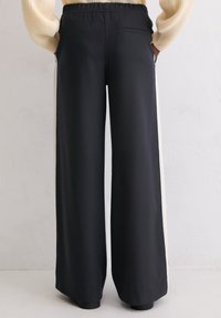 Osoba ubrana w czarne szerokie spodnie z białymi paskami po bokach i kremowy sweter w prążki, stojąca na tle białej teksturowanej ściany.