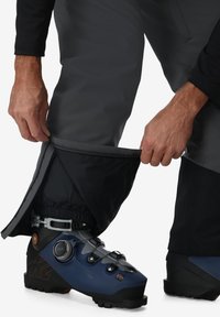 Schwarze Skihosen mit einem Reißverschluss am Unterschenkel, der einen blauen Skischuh mit BOA-Verschlusssystem und grauen Elementen zeigt. Nahaufnahme einer Hand, die anpasst.