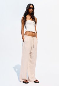 Pantalons larges beiges à côtes légers, avec une taille à cordon de serrage, associés à un haut court blanc avec un ourlet à volants. Lunettes de soleil sombres portées.