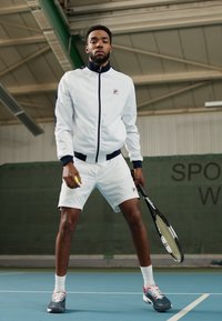 Weißer, zip-fester Sportjacke mit marineblauen Akzenten, kombiniert mit weißen Shorts. Das Model hält einen Tennisschläger und einen Ball und steht auf einem blauen Platz.