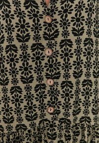 Svart blommönster på ljus olivfärgad tyg, med v-ringning och träknappar. Volangkant ger struktur och detalj.