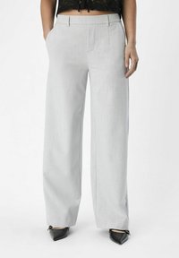 Pantalon large gris clair avec une texture lisse, doté de poches avant et d'une coupe ajustée. Porté avec des chaussures noires à bout pointu.