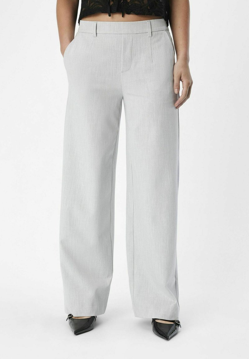 Pantalon large gris clair avec une texture lisse, doté de poches avant et d'une coupe ajustée. Porté avec des chaussures noires à bout pointu.