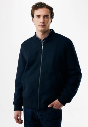 Giovane uomo con capelli ricci che indossa una giacca nera con cerniera e jeans scuri, in piedi con una mano in tasca davanti a uno sfondo semplice.