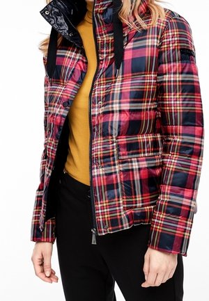 Person trägt eine rot-navy karierte Steppjacke über einem senfgelben Oberteil mit schwarzen Hosen, steht vor weißem Hintergrund.