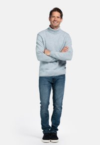 Lichtblauwe, gestructureerde coltrui, gecombineerd met donkerblauwe jeans en zwarte schoenen. Model staat met de armen gekruist tegen een witte achtergrond.