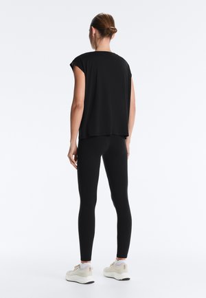 Frau steht mit dem Rücken zur Kamera, trägt ein locker sitzendes schwarzes ärmelloses Oberteil, schwarze Leggings, weiße Socken und beige Turnschuhe.