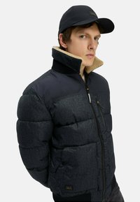 Puffjakke med en marineblå overdel og en mørkegrå underdel, der har en pelsforet krave, lynlåslommer og ribbede manchetter. Sort cap inkluderet.