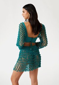 Turquoise haakcover-up met lange mouwen en een opengehaakt ontwerp. Heeft een aansluitend lijfje, verstelbare rugstrik en een franjeszoom.