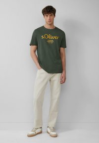 Dunkelgrünes T-Shirt mit kurzen Ärmeln und gelbem Markentext, kombiniert mit hellbeigen Hosen und weißen Sneakers mit braunen Akzenten.