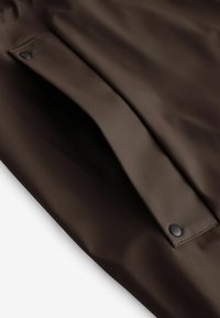 Tessuto impermeabile marrone con una texture liscia. Presenta una tasca con un pulsante a scatto nero sulla patta, con dettagli di cucitura in evidenza.