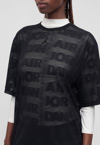 Personne portant un t-shirt en mesh noir Air Jordan par-dessus une chemise blanche à manches longues, affichant des motifs de texte "Air Jordan" répétés.