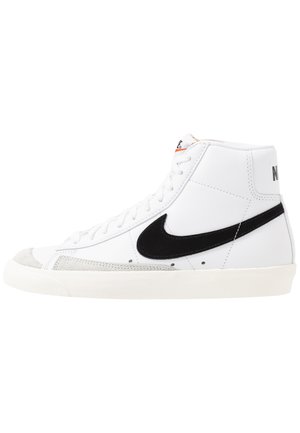Baskets montantes blanches avec des accents noirs, bout en suède, semelle en caoutchouc texturé et couture visible sur une tige en cuir lisse.