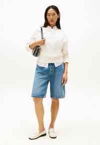 Felpa crema con scollo rotondo, abbinata a pantaloni bermuda in denim blu. Camicia bianca sotto e una borsa a tracolla nera con fantasia. Mocassini bianchi.